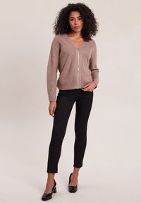 Braune Strickjacke mit V-Ausschnitt und Reißverschluss, mit strukturierten vertikalen Linien. Kombiniert mit schwarzen eng sitzenden Hosen und High Heels.