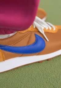Sapatilha Nike castanho-claro com swoosh azul, atacadores brancos e tecido texturizado; posicionada sobre uma superfície verde.