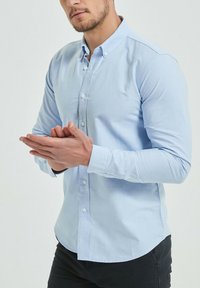 Chemise bleu clair à manches longues et col classique, fabriquée en tissu lisse. Portée avec un pantalon sombre, les mains jointes.