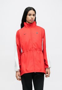 Jachetă roșie de tip windbreaker cu guler înalt, talie elastică și mâneci albe. Prezintă un logo mic pe piept și fermoar frontal.