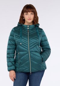 Žena oblečená v tyrkysovém prošívaném péřovém kabátu s kapucí, se zipovými kapsami a dvěma stříbrnými knoflíky na límci, v kombinaci s modrými džínami.