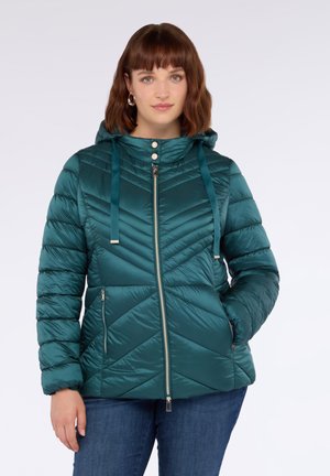 Frau trägt eine türkisfarbene, gesteppte, kapuzenlose Daunenjacke mit Reißverschlusstaschen und zwei silbernen Druckknöpfen am Kragen, kombiniert mit blauen Jeans.