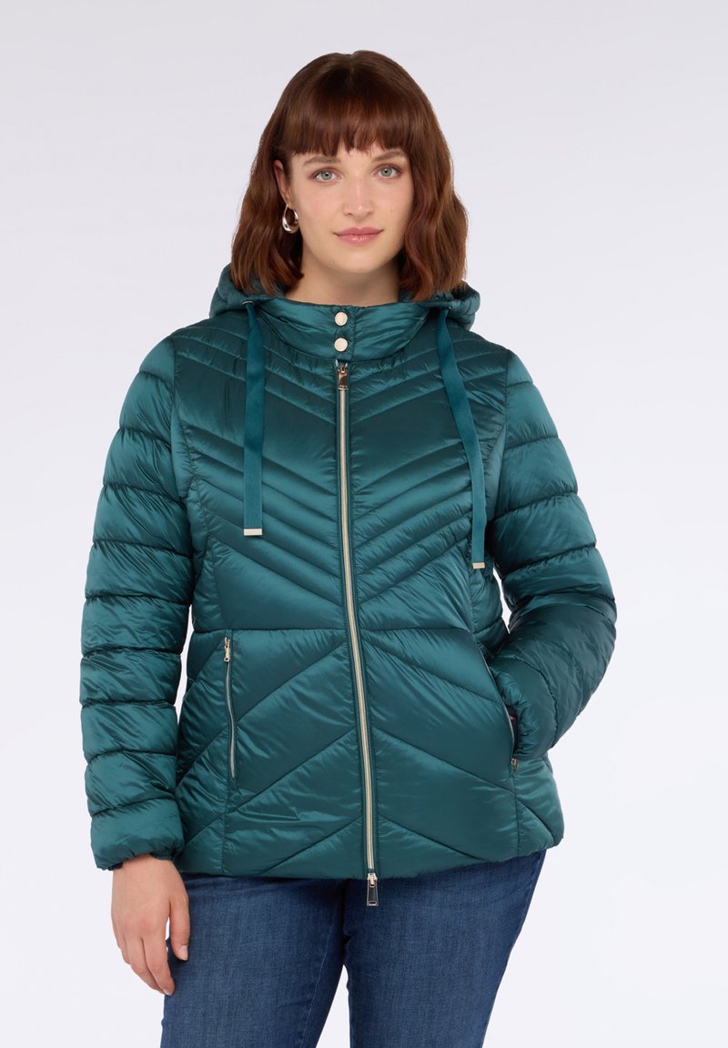 Žena oblečená v tyrkysovém prošívaném péřovém kabátu s kapucí, se zipovými kapsami a dvěma stříbrnými knoflíky na límci, v kombinaci s modrými džínami.