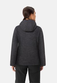 Vrouw met donker haar in een staart, gekleed in een donkergrijze, gestructureerde hoodie en zwarte broek, gezien van achteren tegen een effen achtergrond.