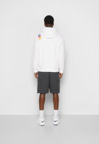 Man som står med ryggen mot kameran, iförd en vit huvtröja med tre färgade Nike-loggor på axeln, mörkgrå cargoshorts, vita strumpor och vita Nike-skor.