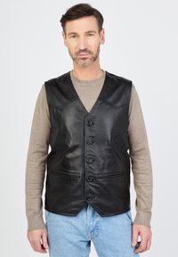 Schwarze Lederweste mit V-Ausschnitt, fünf Knöpfen und zwei vorderen Taschen. Sie hat eine glatte Textur und eine taillierte Passform. Getragen über einem beigen Pullover.