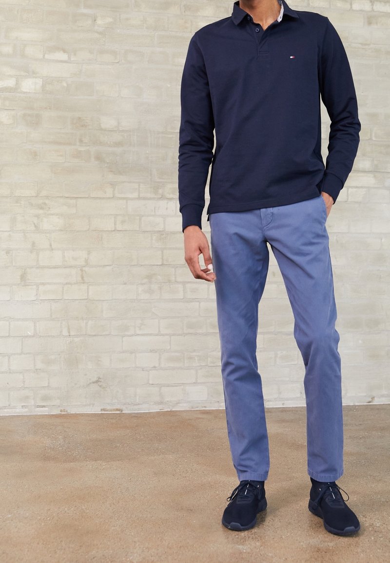 Homme portant un polo à manches longues bleu marine, un pantalon chino bleu clair et des baskets bleu foncé, debout contre un mur en briques clair.