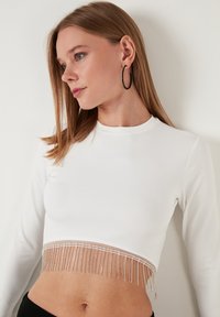 Bjela dugih rukava kratka majica s prozirnim rubom, ukrašena metalnim perlicama. Glatka tkanina, prilagođeni kroj i minimalistički okrugli izrez.