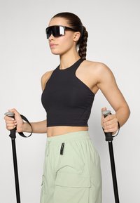 Mustmustega crop-top, helekroonilised cargo-püksid ja suured mustad prillid. Mudel hoiab trekkingupulte, millel on hallid kummist käepidemed.