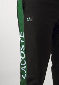 Pantaloni atletici negri cu un panou lateral verde, având inscripția "LACOSTE" în litere albe și un mic logo de crocodil brodat deasupra.