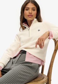 Cremfarbener Pullover mit Reißverschluss-Kragen, besticktem Logo, getragen über einem rosa Hemd mit Kragen. Graue Nadelstreifenhose und ein Hintergrund mit einem geflochtenen Stuhl.