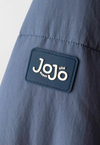 Prim-plan al unei etichete din material albastru pe o îmbrăcăminte, cu "JoJo maman bébé" brodat în litere albe pe un material texturat de culoare albastră.