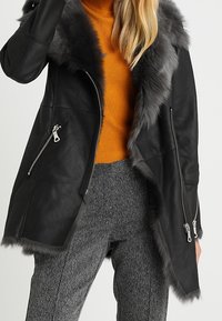 Sort læderjakke med grå falsk pelskrave; har sølv lynlåse på siderne. Båret over en orange sweater og grå mønstrede bukser.