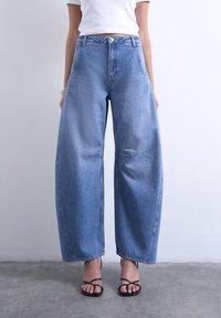 Άτομο που φοράει μπλε τζιν παντελόνι με ψηλή μέση και φαρδιά πόδια, λευκό κοντό τοπ, ασημένιο βραχιόλι και μαύρα σανδάλια με τακούνια και λουριά, στέκεται σε γκρι πάτωμα.