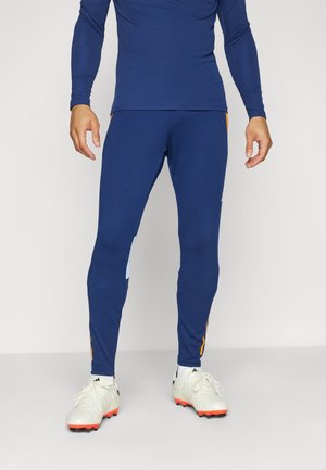 Mann trägt blaue Sportleggings und weiße Stollenschuhe mit roten Sohlen, steht vor einem einfarbigen hellen Hintergrund.