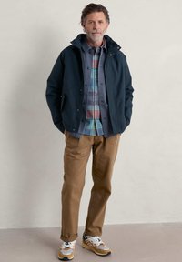 Chaqueta impermeable navy sobre una camisa de cuadros gris, pantalones beige y zapatillas coloridas. La chaqueta cuenta con una capucha con cierres de botón.
