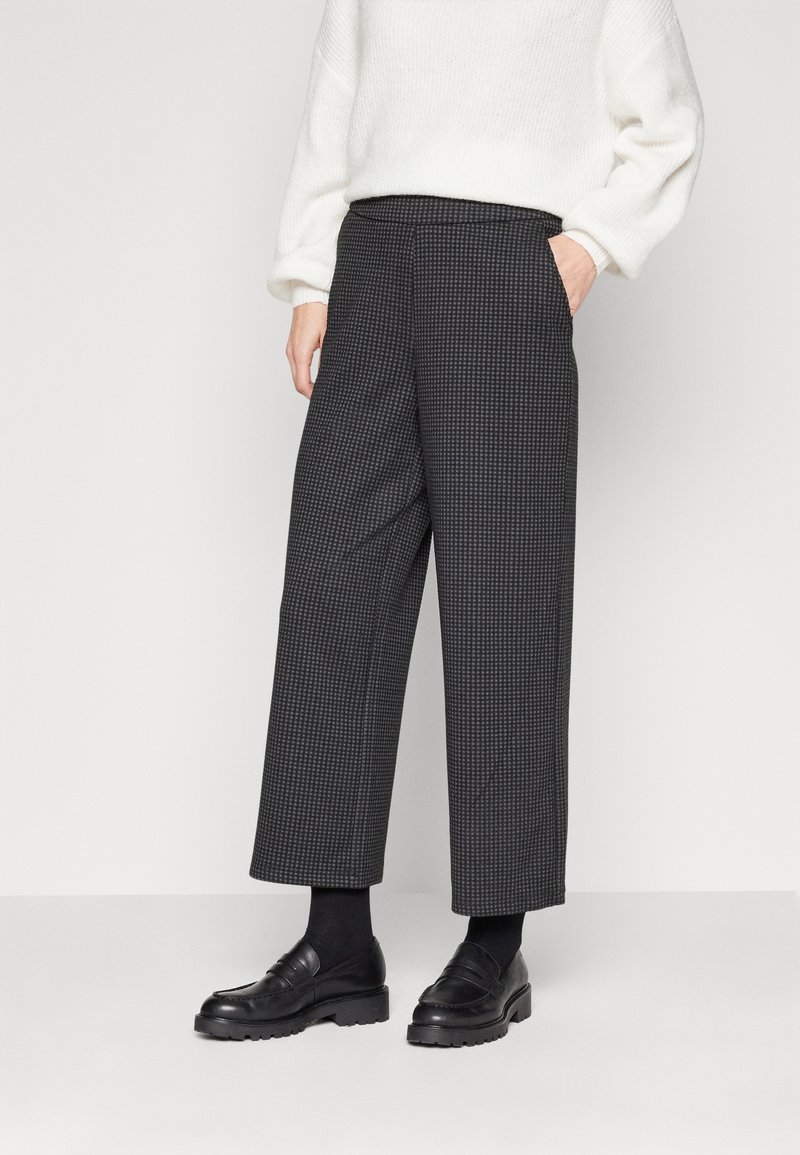Personne portant un pantalon large à carreaux noirs et gris raccourci, des chaussettes noires, des mocassins noirs et un pull blanc à manches longues, debout contre un mur blanc.