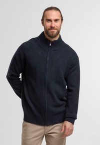 Marinefarbener Reißverschluss-Pullover mit geripptem Stehkragen, aus weichem Strickmaterial, mit einem Frontreißverschluss und langen Ärmeln. Über hellen Hosen getragen.