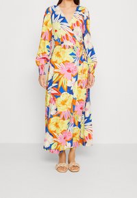 Vestido largo estilo envolvente con un vibrante estampado floral en rosa, amarillo y azul, con mangas largas y cintura anudada. Combinado con sandalias ligeras.