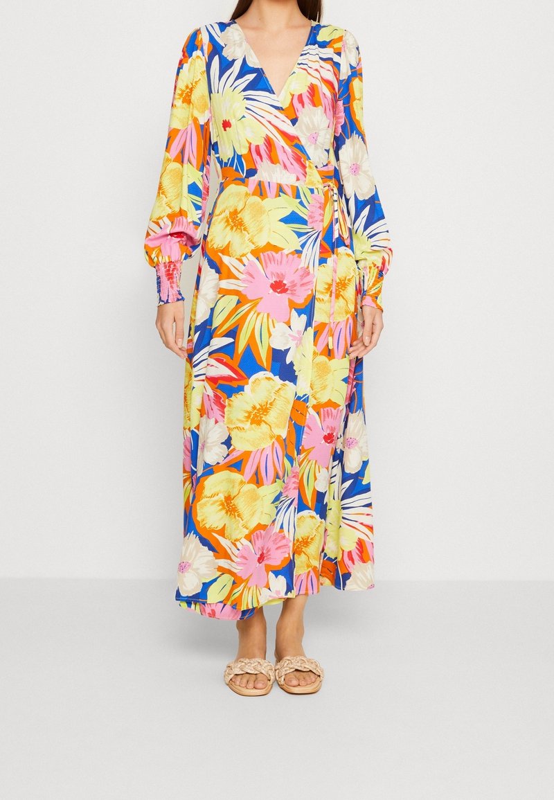 Vestido largo estilo envolvente con un vibrante estampado floral en rosa, amarillo y azul, con mangas largas y cintura anudada. Combinado con sandalias ligeras.