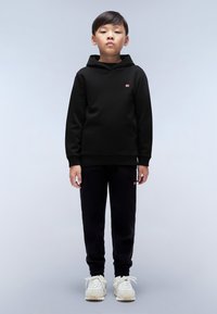Felpa nera con un piccolo logo rosso sul petto, abbinata a pantaloni jogger neri e sneakers di colore chiaro. Design semplice, vestibilità rilassata.