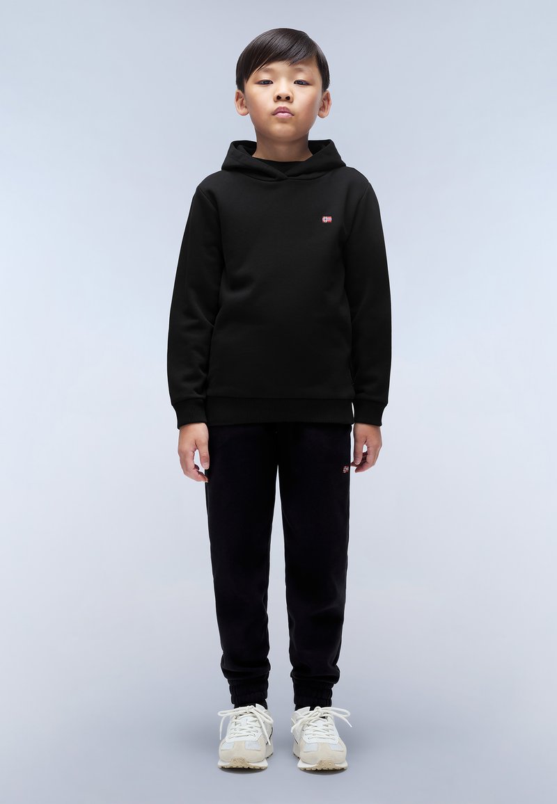 Felpa nera con un piccolo logo rosso sul petto, abbinata a pantaloni jogger neri e sneakers di colore chiaro. Design semplice, vestibilità rilassata.
