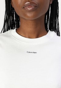 Hvit bomull T-skjorte med rund hals, med en liten svart "Calvin Klein" logo på brystet. Glatt tekstur.