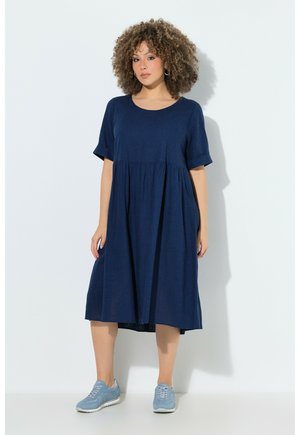 Femme aux cheveux bouclés portant une robe midi bleu marine ample et des baskets bleu clair, debout sur un fond blanc uni.