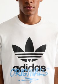 Maglietta bianca con un grande logo Adidas nero che presenta tre strisce e un testo stilizzato blu "ORIGINALS" in basso.