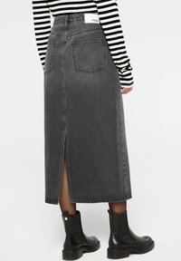 Schwarzer Denim-Midi-Rock mit einem Schlitz hinten, zwei hinteren Taschen, einem Etikett am Bund und geradem Schnitt. Kombiniert mit schwarzen Stiefeletten.