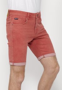 Pantalones cortos de mezclilla rojos con un corte ajustado, que cuentan con dobladillos enrollados, bolsillos frontales y una cremallera. La tela tiene una ligera textura desgastada.