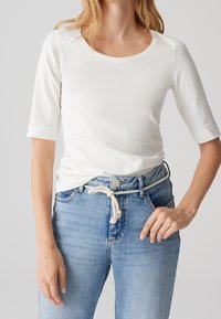 Femme portant un haut blanc à manches aux coudes rentré dans un jean bleu clair avec une ceinture en corde blanche nouée à la taille.