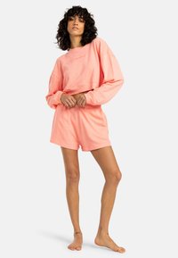 Loungewear set v barvni koralni, ki vključuje skrajšano pulover z dolgimi rokavi in kratke hlače, oboje iz mehkega, lahkotnega materiala.