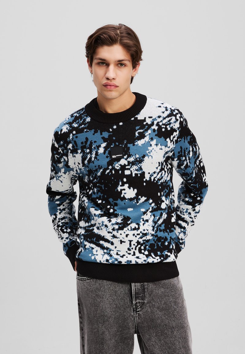 Blå, sort og hvid mønstret sweater med ribbet sort halsudskæring og ærmekanter, lavet af tekstureret stof. Bæres med grå jeans.