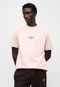 Ανοιχτό ροζ κοντομάνικη μπλούζα με στρογγυλή λαιμόκοψη και πράσινο λογότυπο "Originals". Διαθέτει ραγκλάν μανίκια και απαλό βαμβακερό ύφασμα.