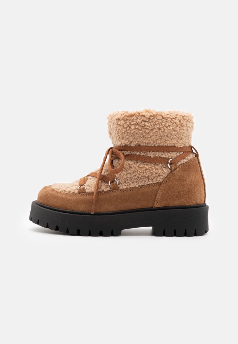 Bottine en suede beige avec une doublure en fourrure crème, des lacets marron, des accessoires en métal argenté, et une épaisse semelle en caoutchouc noir avec un motif de semelle robuste.