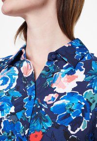 Camicia floreale in blu navy con grandi fiori blu, rosa e verdi. Presenta un colletto con bottoni e una texture di tessuto liscia.