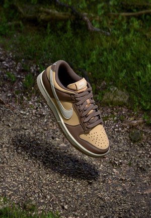 Brązowy i beżowy sneaker Nike z białymi sznurówkami w kształcie znaku Swoosh, zawieszony nad żwirową ścieżką z zieloną trawą w tle.