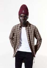 Bruin en beige geblokt overhemd, wit T-shirt eronder, zwarte corduroy broek en een bordeauxrode muts met een logo aan de voorkant. Effen achtergrond.