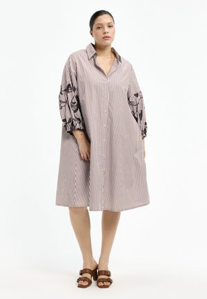 Frau trägt ein locker sitzendes, knielanges, gestreiftes Kleid mit Knöpfen und dreiviertel Ärmeln, bestickt mit schwarzen Blumendesigns, kombiniert mit braunen Sandalen mit Absatz.