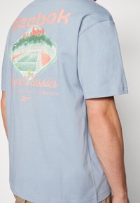 Světle modré bavlněné tričko s grafickým designem tenisového zápasu a panoramatu na zadní straně, s růžovým textem a akcenty.