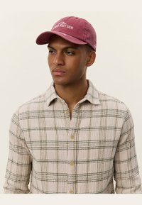 Maroon cotton baseball cap with "Upper West Side" embroidered in white; worn with a beige and green checkered flannel shirt.