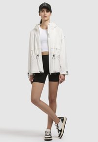 Jachetă albă impermeabilă cu glugă, accente negre, talie cu șnur și două buzunare frontale. Asortată cu un top alb cropped și pantaloni scurți negri.