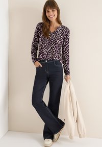 Paarse top met lange mouwen en dierenprint, gecombineerd met donkerblauwe spijkerbroek met hoge taille, en een crèmekleurige jas over de arm.