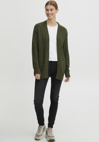 Cardigan olive, à devant ouvert, avec des poignets et un ourlet côtelés, porté par-dessus un t-shirt blanc et associé à un jean slim noir et des baskets beiges.
