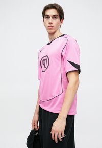 Ροζ αθλητική μπλούζα από διχτυωτό υλικό με μαύρες λεπτομέρειες, που διαθέτει στρογγυλή λαιμόκοψη και κοντά μανίκια. Το μοντέλο κρατάει ένα μαύρο καπέλο.