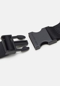 Černý nylonový pásek s plastovou sponou s patentním uzávěrem. Spona je ražena s nápisem "Calvin Klein Jeans." Odolný a funkční design.