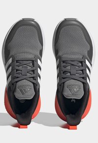 Zapatillas deportivas grises con parte superior de malla, tres franjas blancas, acentos en negro, suela de goma roja y tira de agarre; puntera redondeada y soporte acolchado para el tobillo.