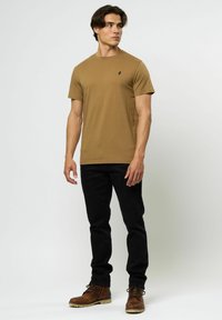 Brun bomulls t-skjorte med korte ermer og en svart logo på brystet, kombinert med svarte jeans og brune snørebånd støvler.