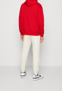 Sudadera con capucha roja y rayas verticales, combinada con pantalones de chándal crema con rayas laterales y zapatillas en blanco y negro.